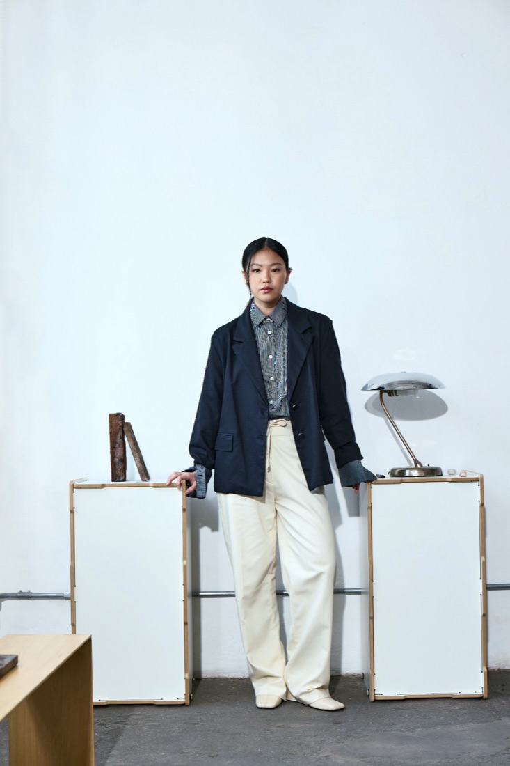 A woman in a navy blazer, striped shirt and cream trousers between two white plinths and a chrome lamp